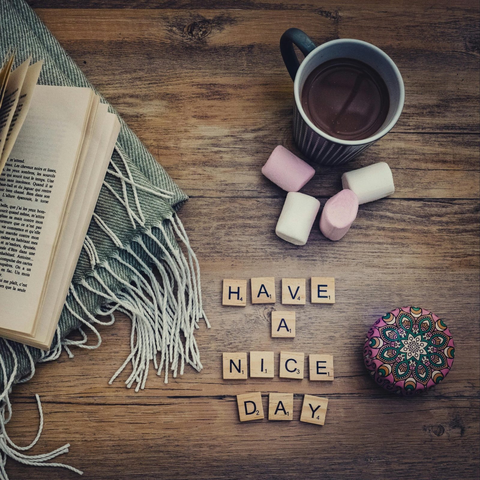 A cozy setup with a coffee, marshmallows, and 'Have a Nice Day' tiles on a wooden table.
